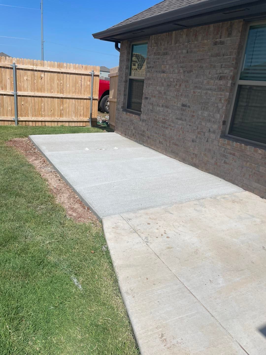 A newly poured concrete patio extends from a brick house wall toward a wooden fence in a grassy backyard.