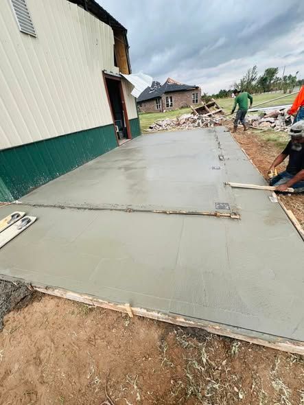 Two people smooth freshly poured concrete for a driveway next to a building.