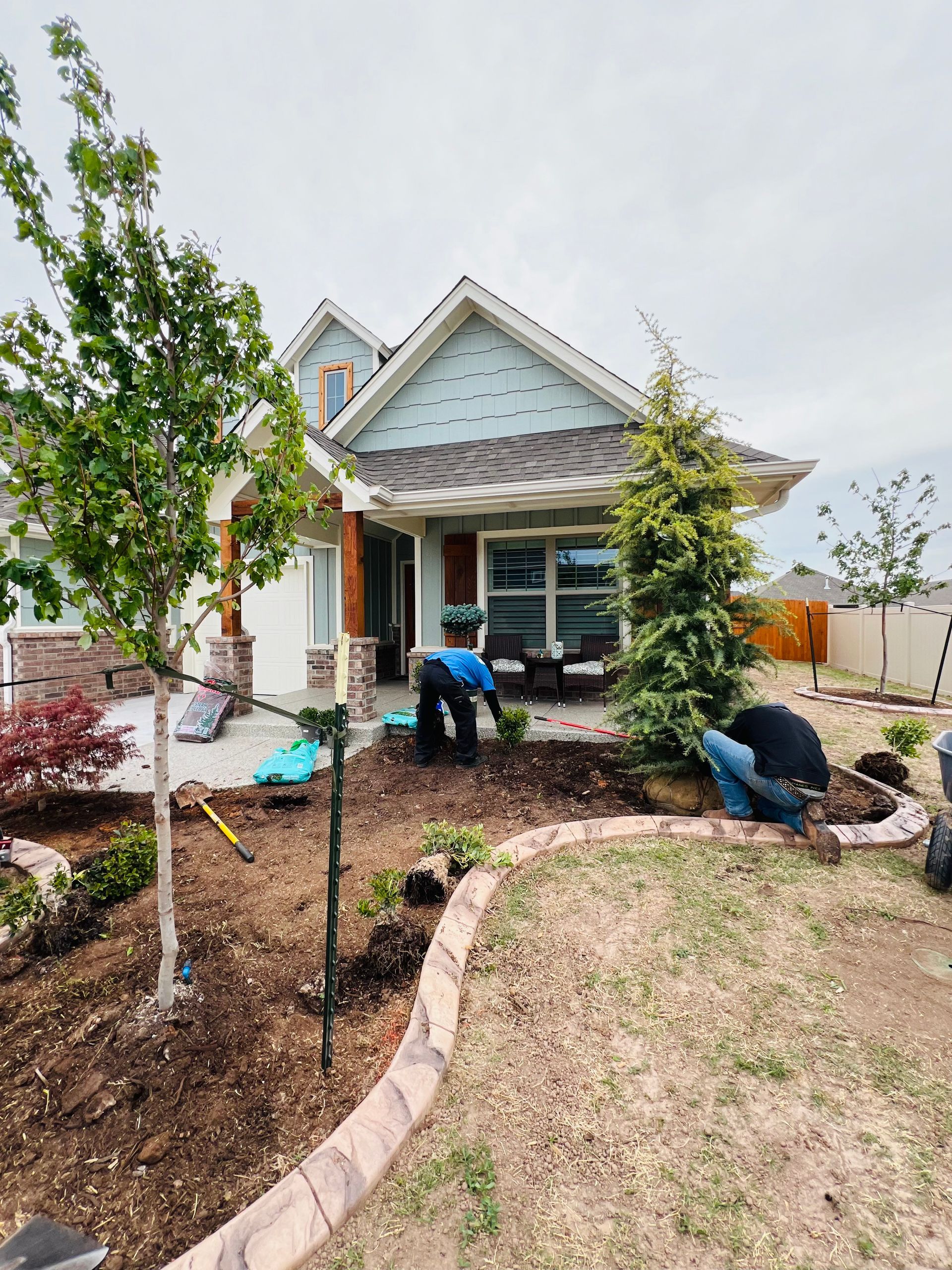 Two people landscaping the front yard of a suburban house with a brick and light blue exterior.