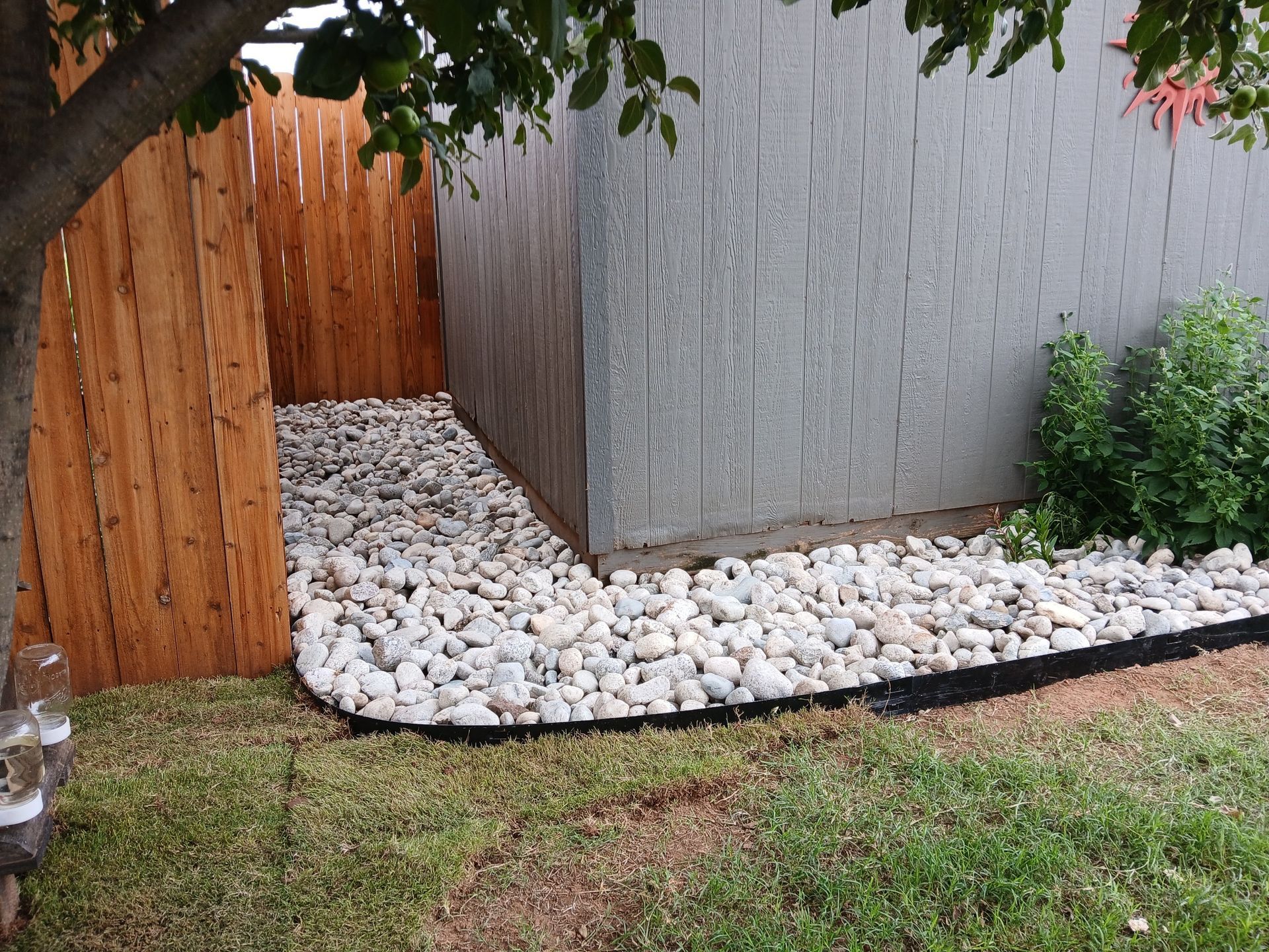A bed of white landscaping rocks borders a gray shed next to a wooden fence and a grassy yard.