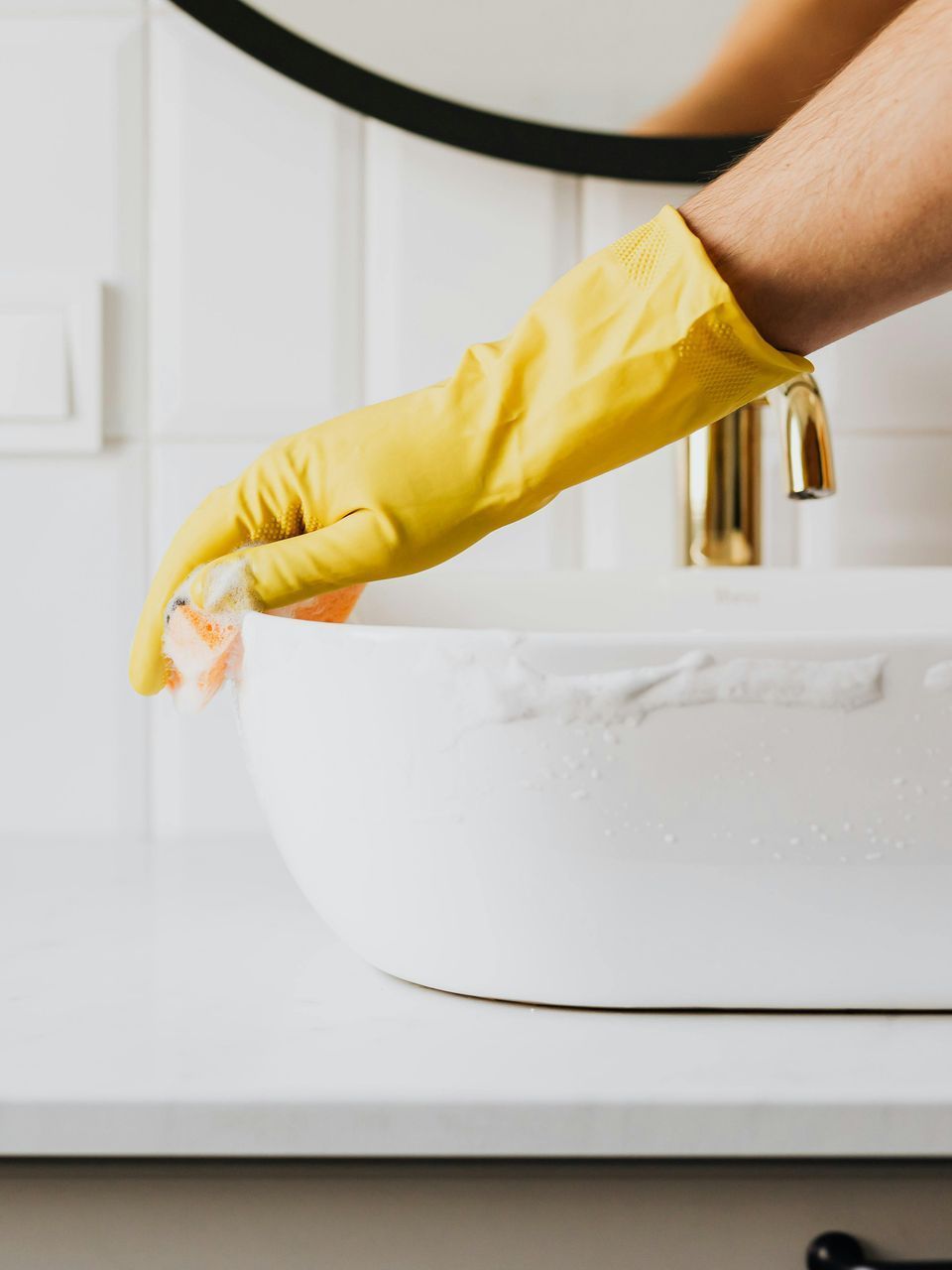 A person wearing a yellow rubber glove is cleaning a bathroom sink.