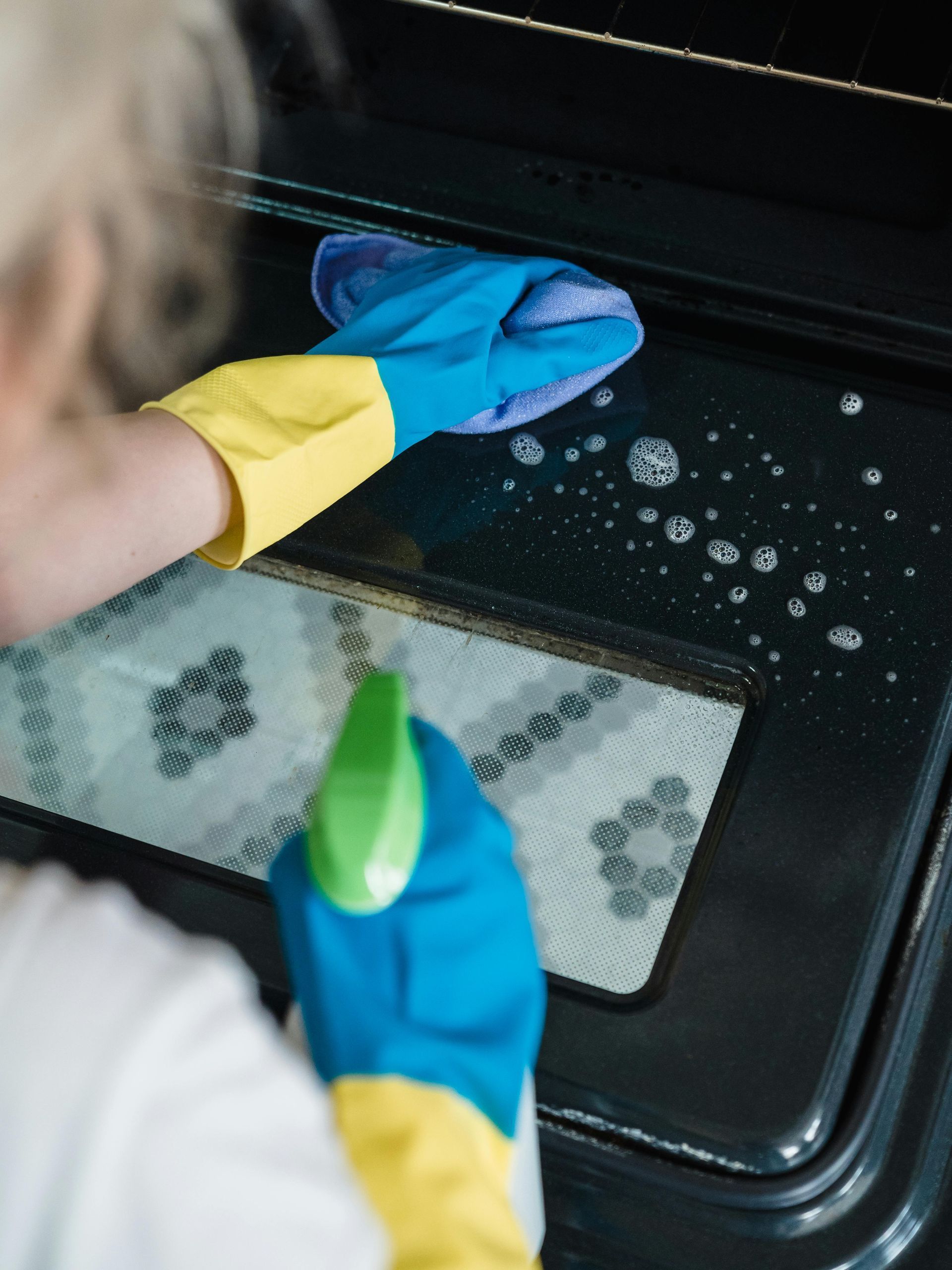 A person wearing rubber gloves is cleaning an oven with a cloth and spray bottle.