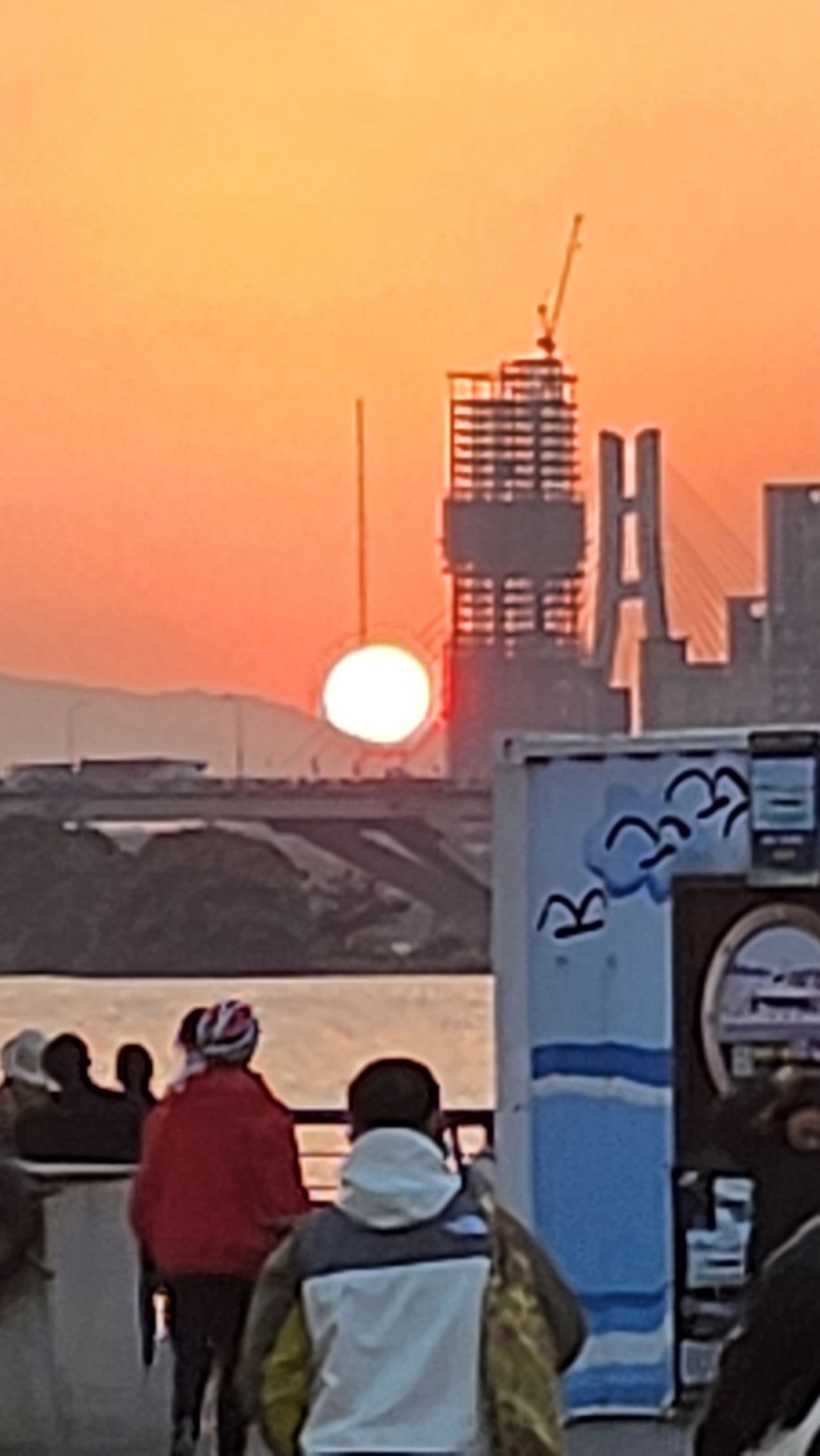 Setting sun over a river, bridge, and construction site; people watching.