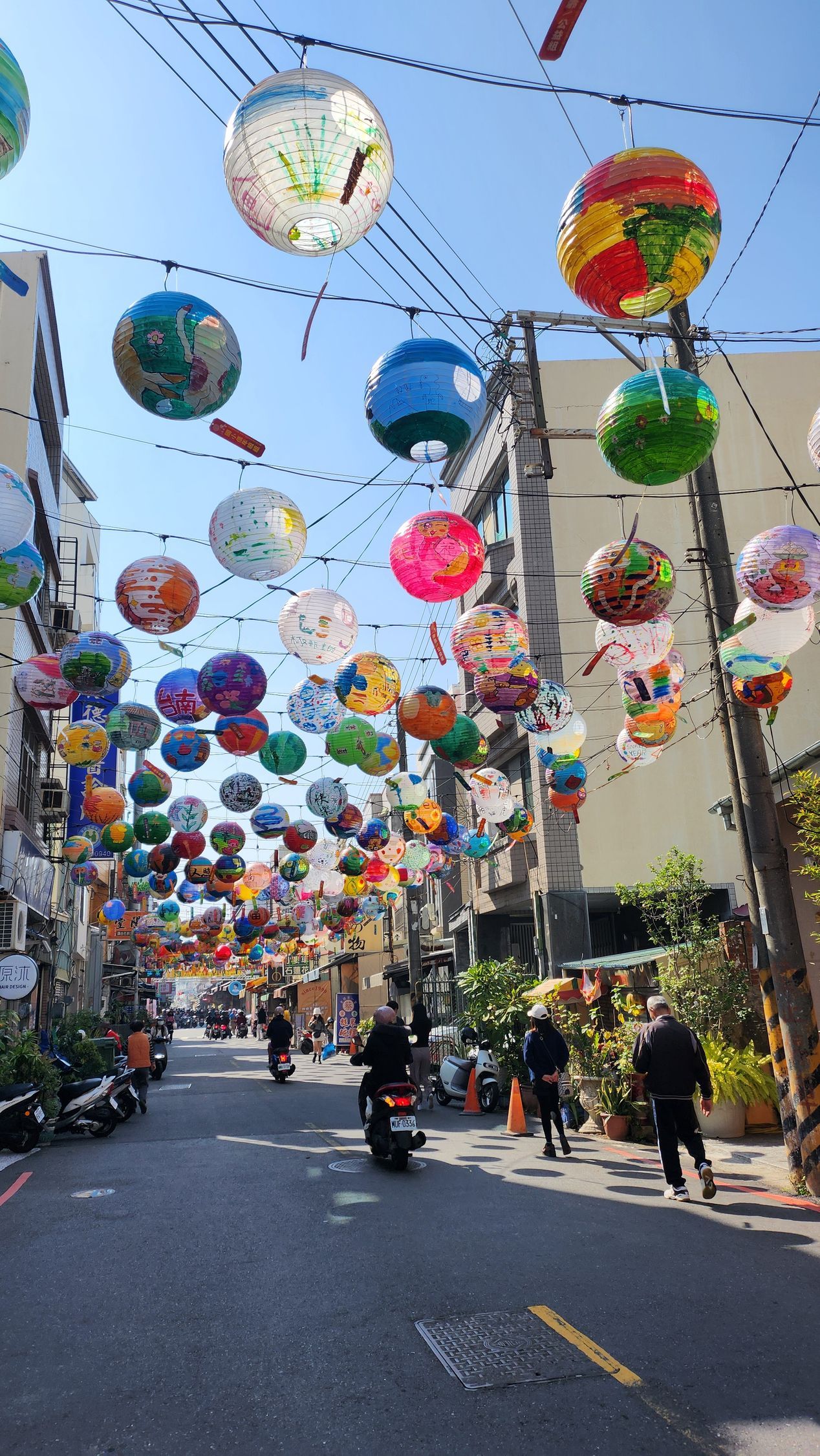 Street decorated with colorful lanterns strung overhead; people and scooters on the street.
