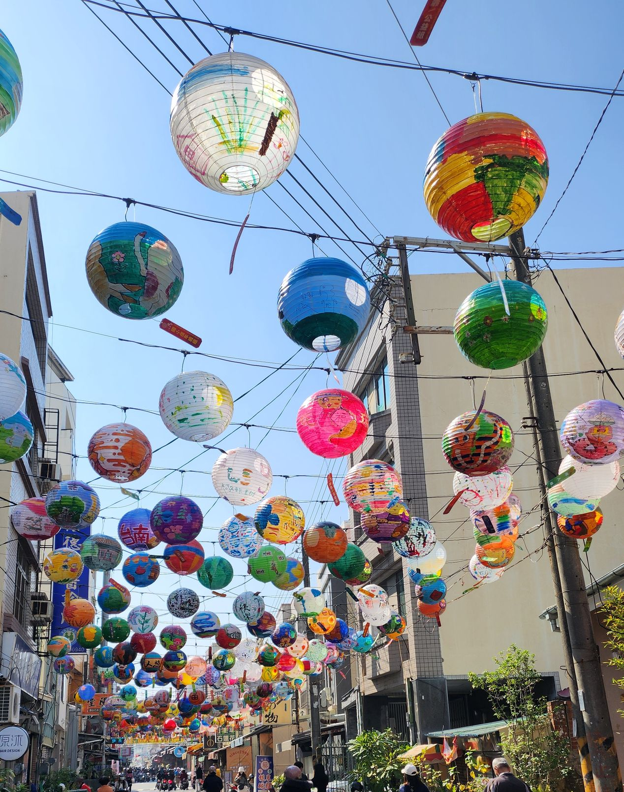 Street decorated with colorful, round lanterns hanging overhead. Bright blue sky and buildings in the background.