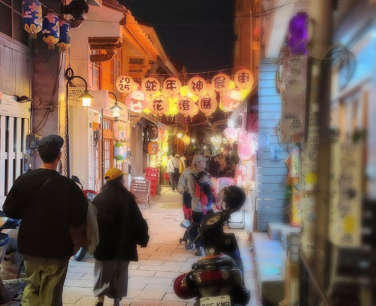 People stroll down a brightly lit alley at night, lined with shops and paper lanterns.