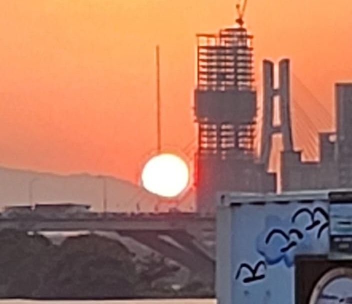 Orange sunset over a cityscape; sun visible, bridge and building silhouettes in the distance.