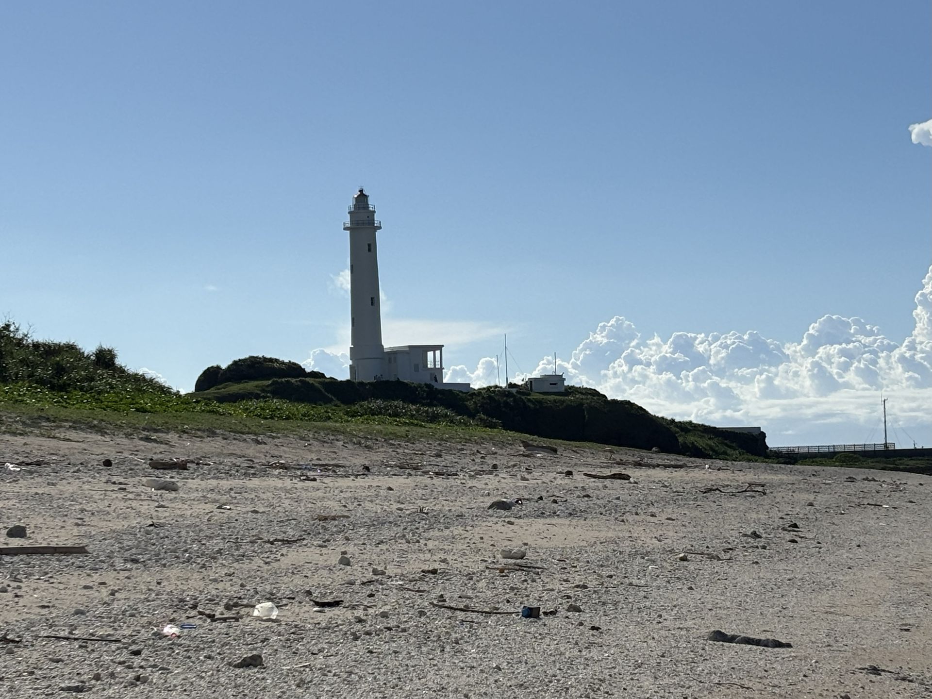 White lighthouse on a hilltop, with low vegetation in front, blue sky with clouds above.