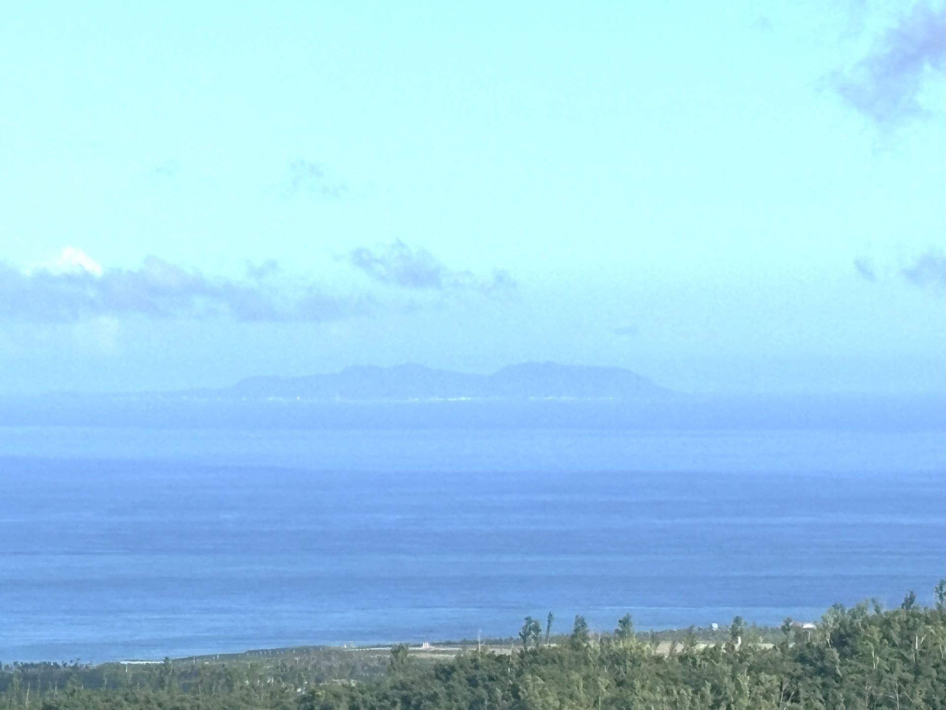 Ocean view with a distant island on the horizon, under a light blue sky, with trees in the foreground.