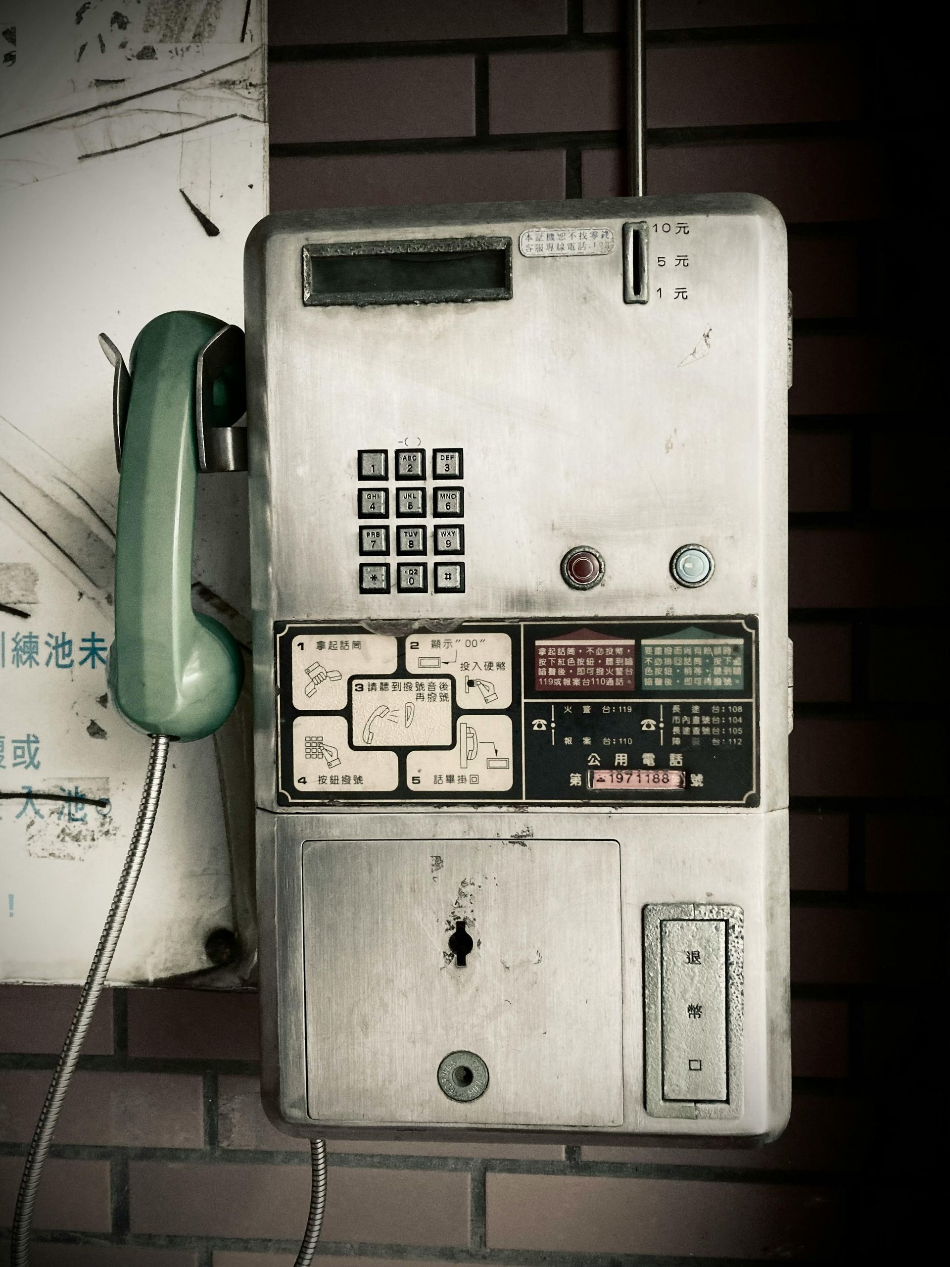 Old, weathered public payphone mounted on a brick wall; green handset, metal body.