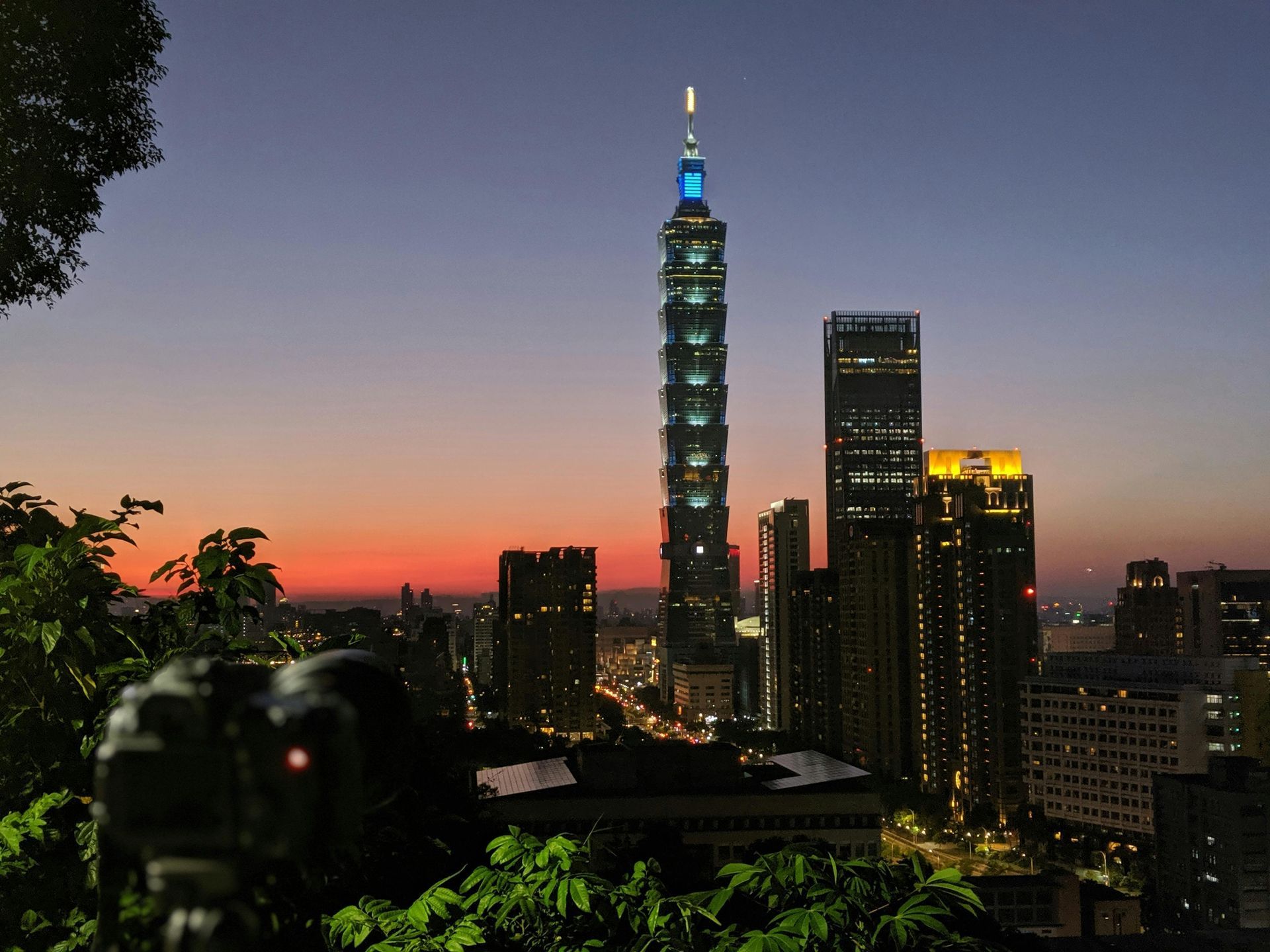 Taipei 101 tower at dusk, seen from a hillside. Skyline includes other lit buildings and a camera in the foreground.