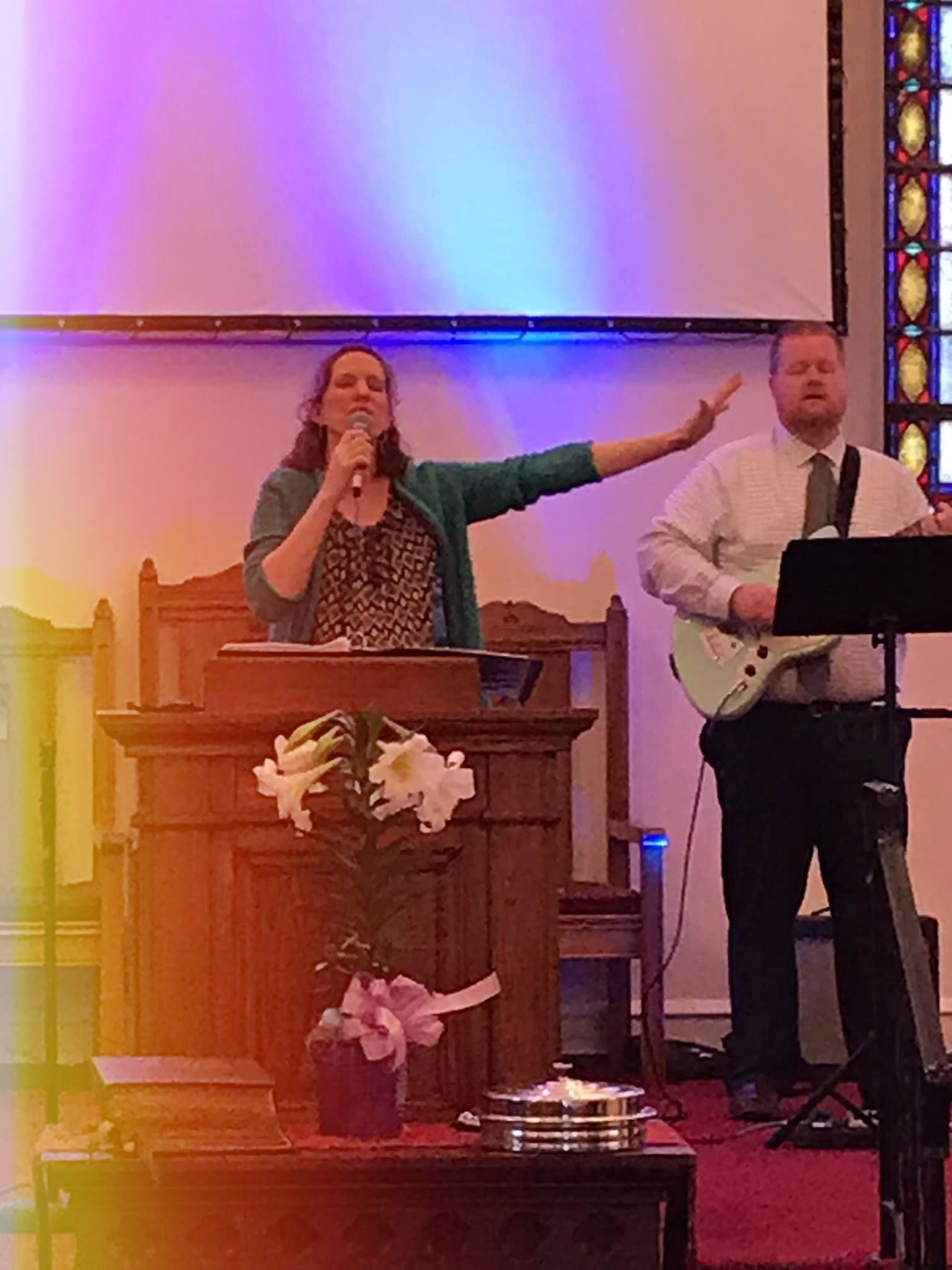 Woman singing at a podium, arm raised. Man plays guitar. Church interior, white screen backdrop, colorful light.