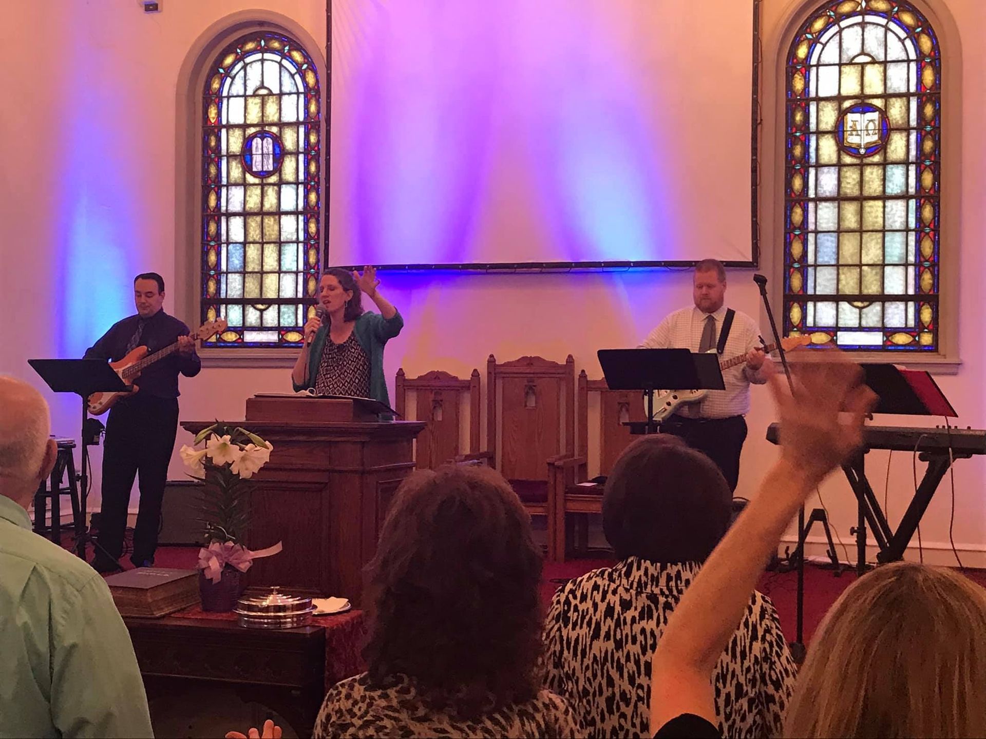 Worship service: a band and singer on stage, people raising hands. Church interior, stained glass windows, purple backdrop.