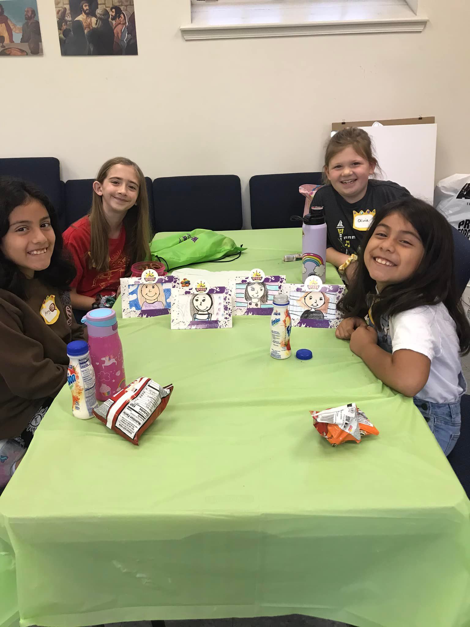 Four girls smile at a table with crafts, snacks, and drinks. Green tablecloth, light-filled room.