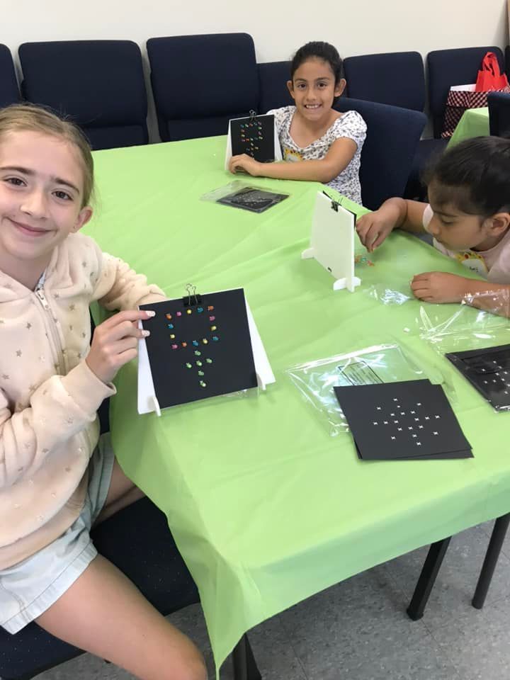 Three girls at a table with craft projects, smiling. One girl holds a black square with colorful dots.