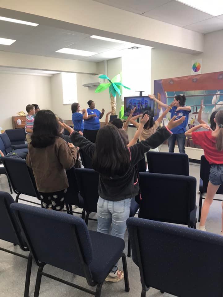 Children with raised arms in front of a stage. Adults in blue shirts lead; palm tree and mural in background.
