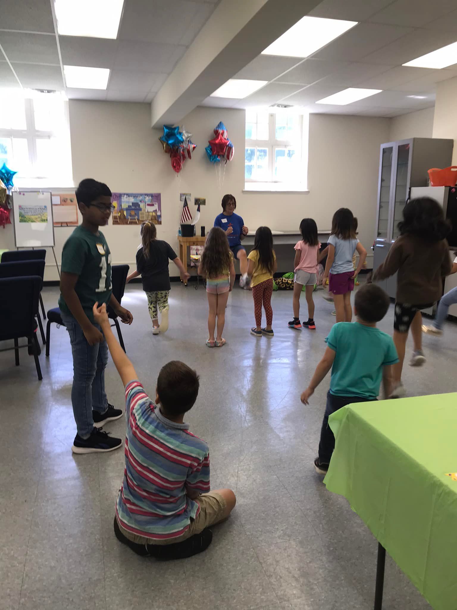 Children dancing in a room, one boy sits and points. Others stand, a facilitator stands in the middle.