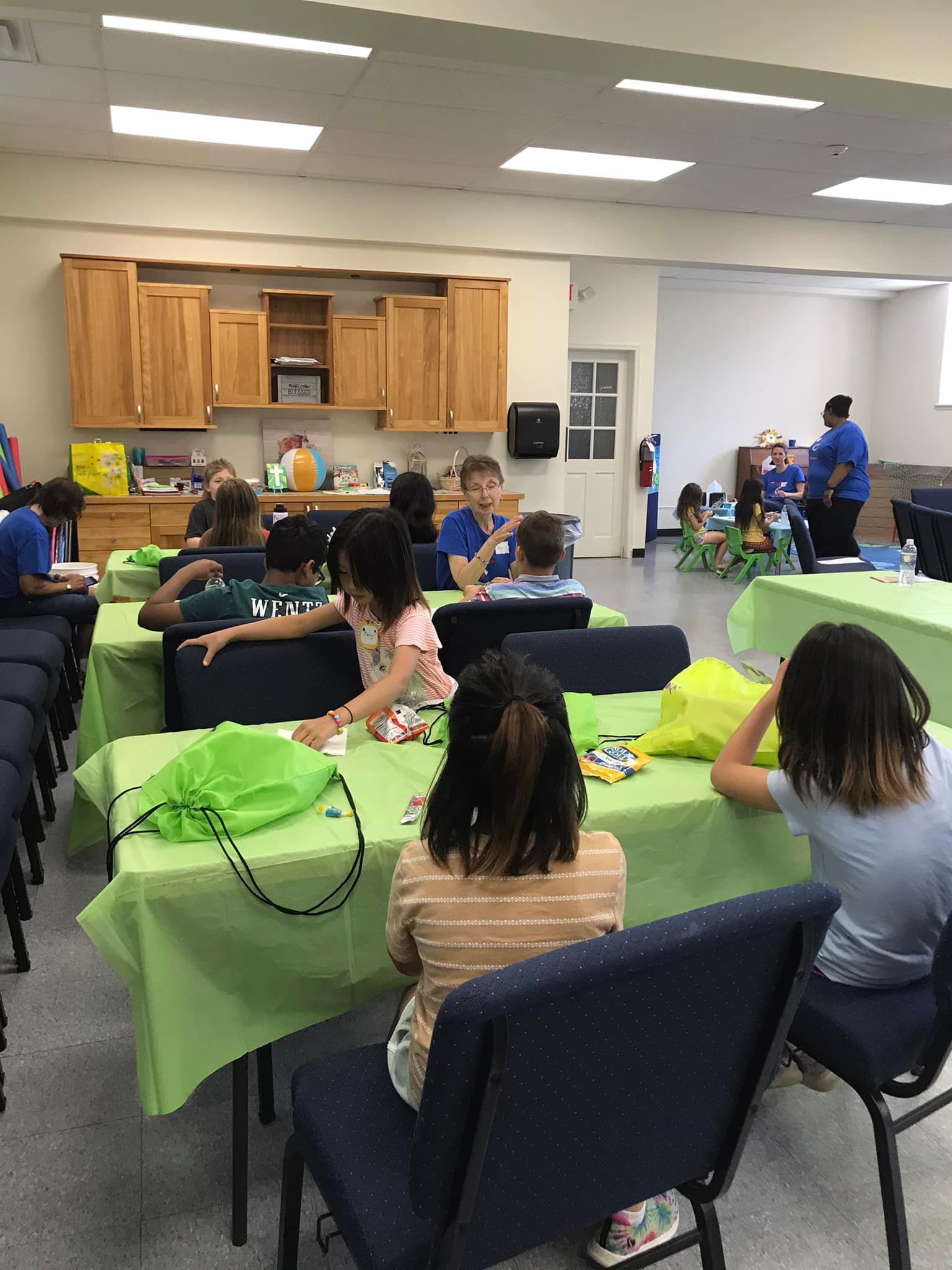 Children at tables in a room with an instructor. Green table cloths and wooden cabinets are visible.