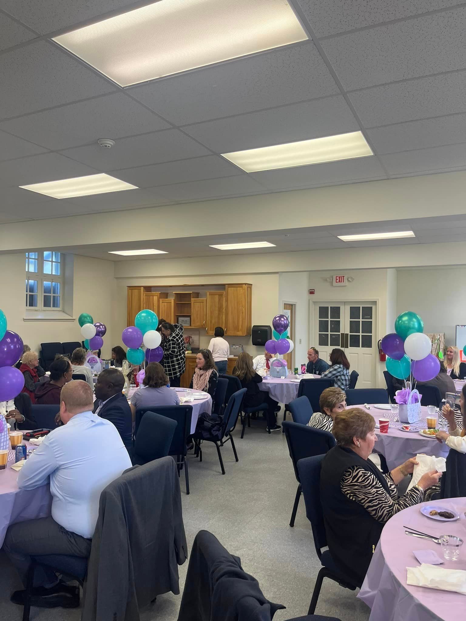 A social gathering in a large room. People seated at round tables decorated with balloons and purple cloths.