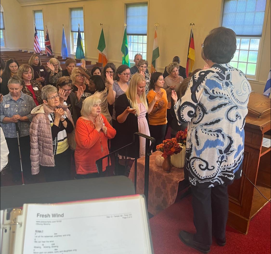 People in church, many with hands raised. Flags behind them, woman in front.