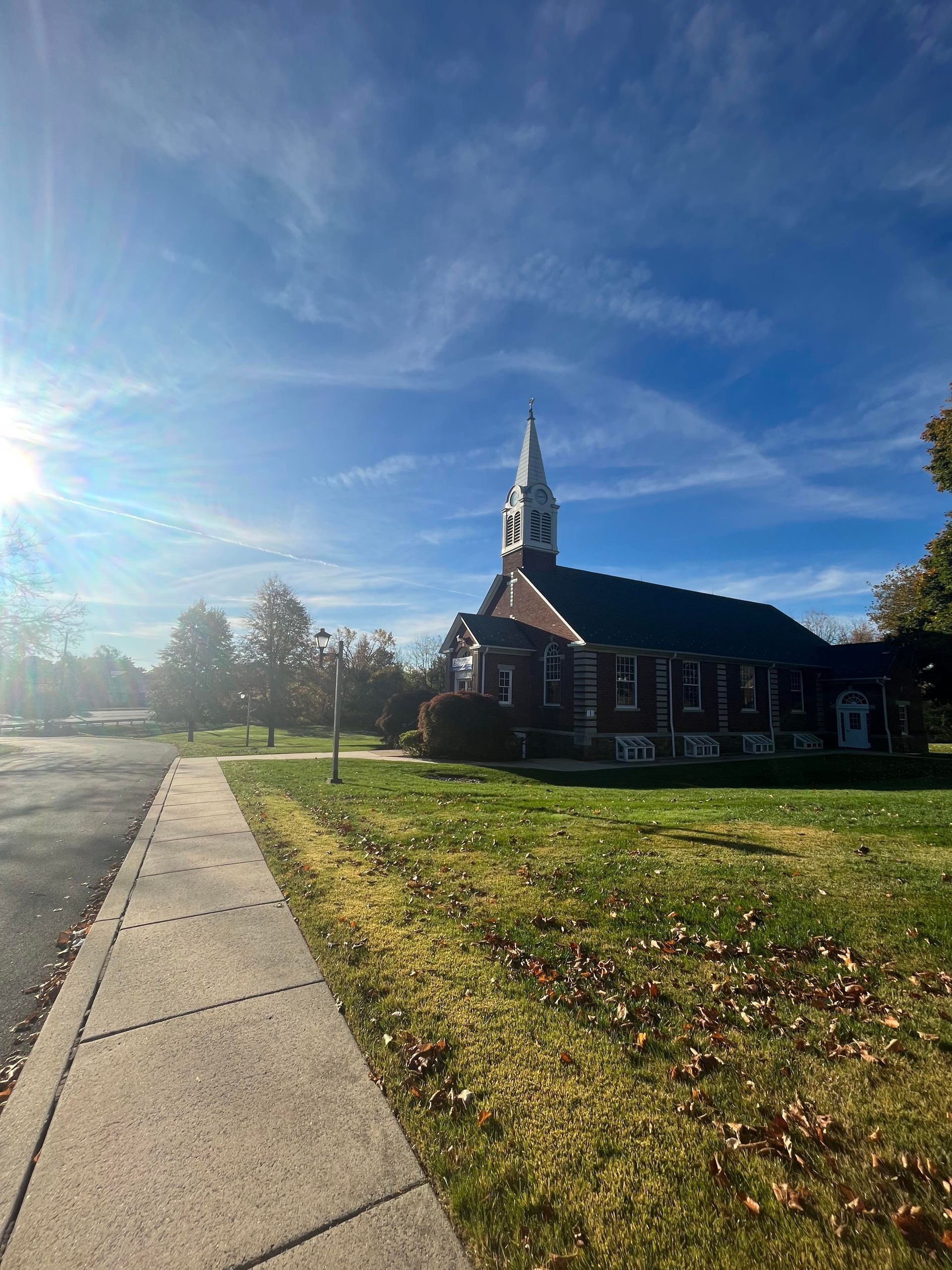 Church building with steeple on a sunny day. Sidewalk in foreground, trees and grass surround.