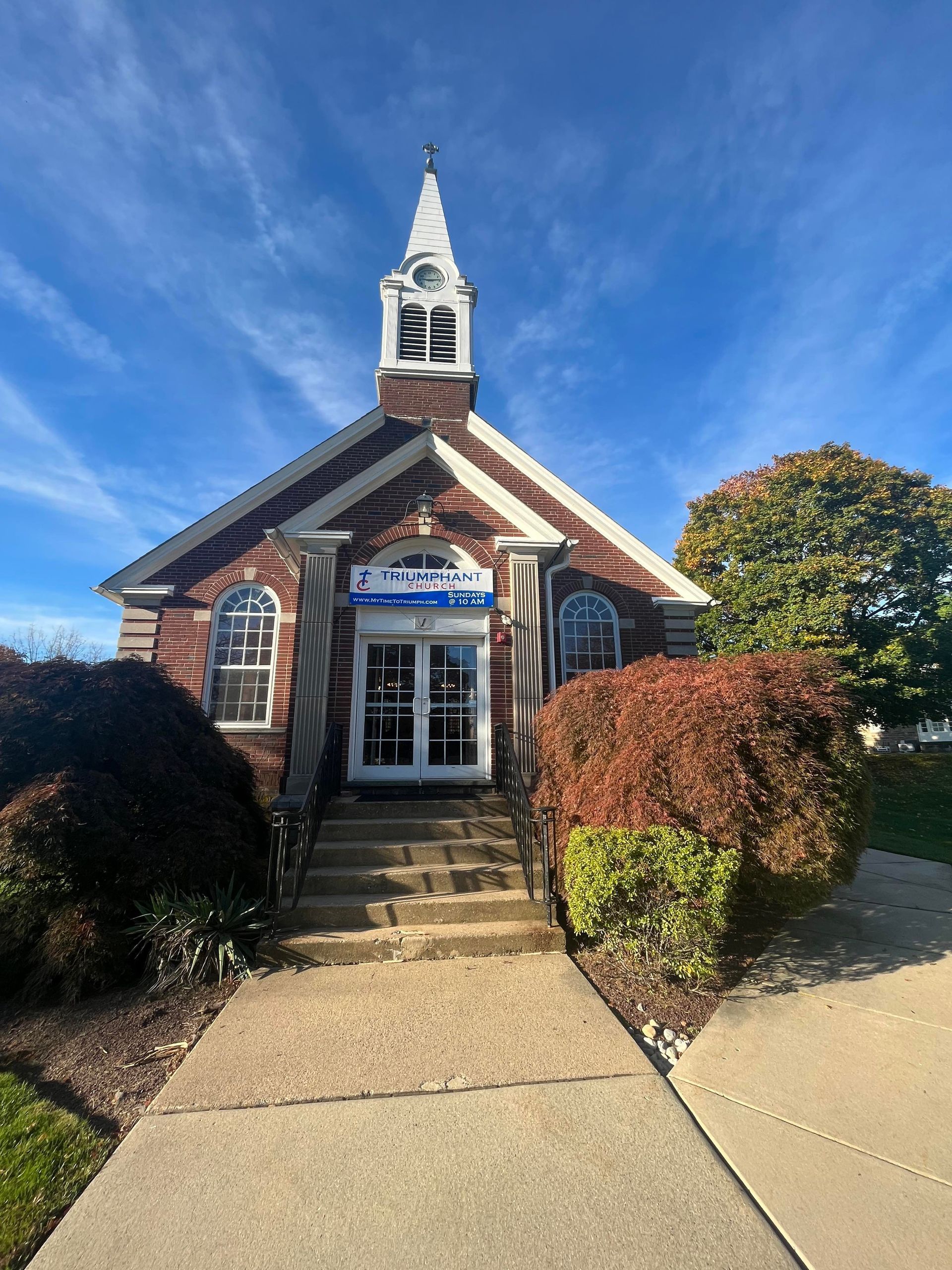 Small red brick building with steeple and shrubs. Blue sky.