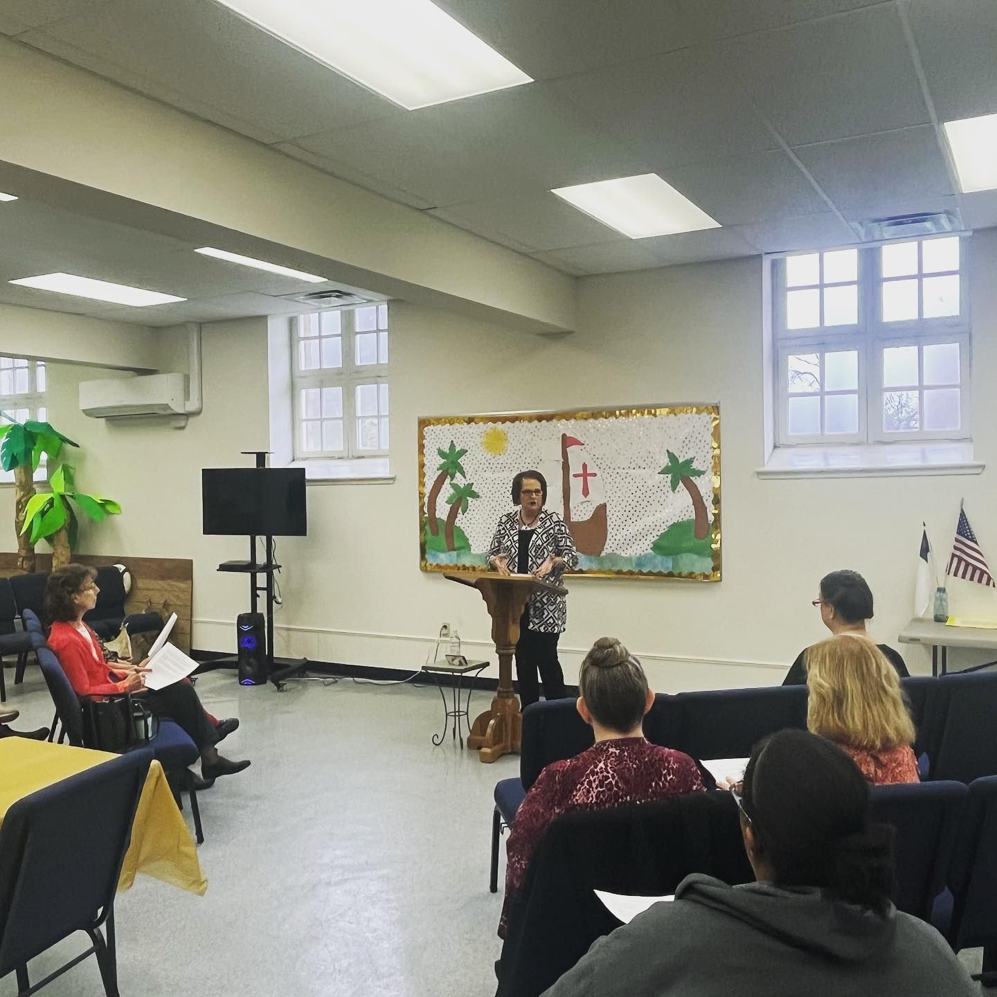 Woman speaking at a podium in a room; audience seated. Decorated backdrop of a ship; several windows.