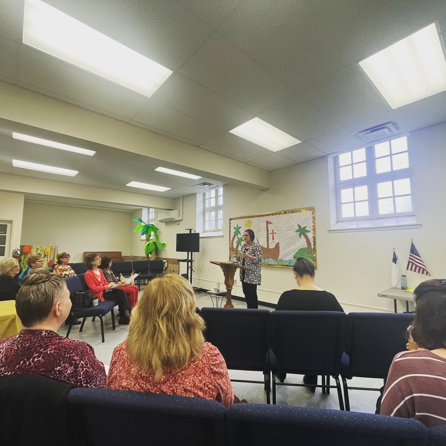 Woman speaking at a podium in a church, audience seated. Beige walls, bright lights, and an American flag.