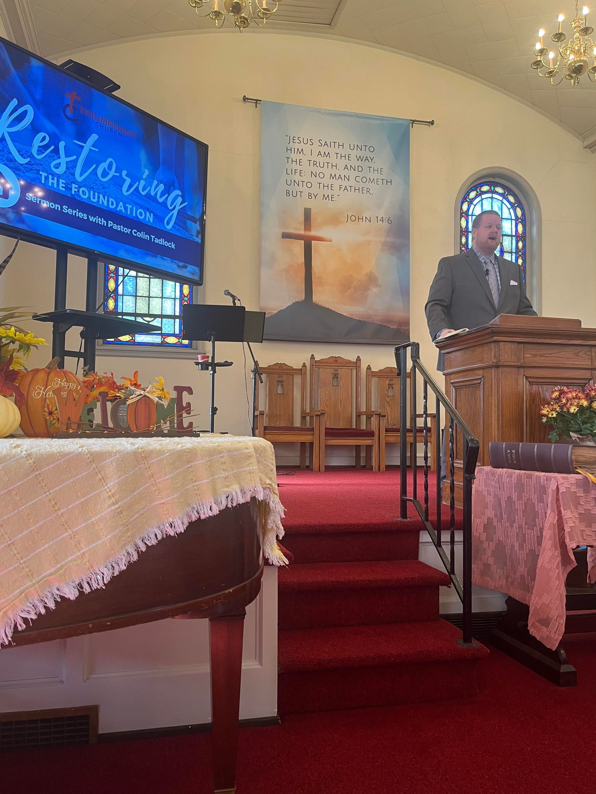 A man in a suit preaches from a pulpit inside a church. A screen displays text
