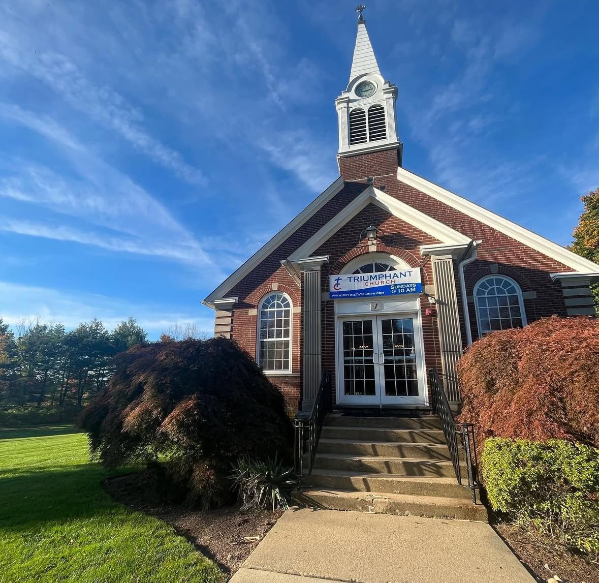 Church with brick facade, steeple, arched windows, and small entrance steps under a blue sky.