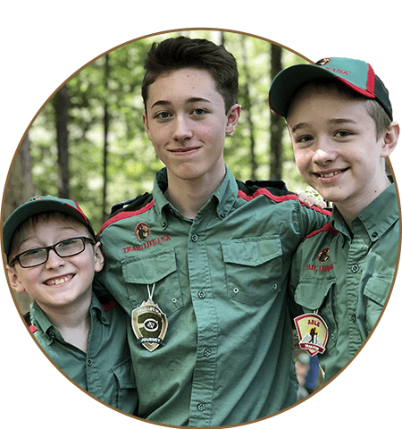 Three smiling Boy Scouts in green uniforms, standing in a wooded area.