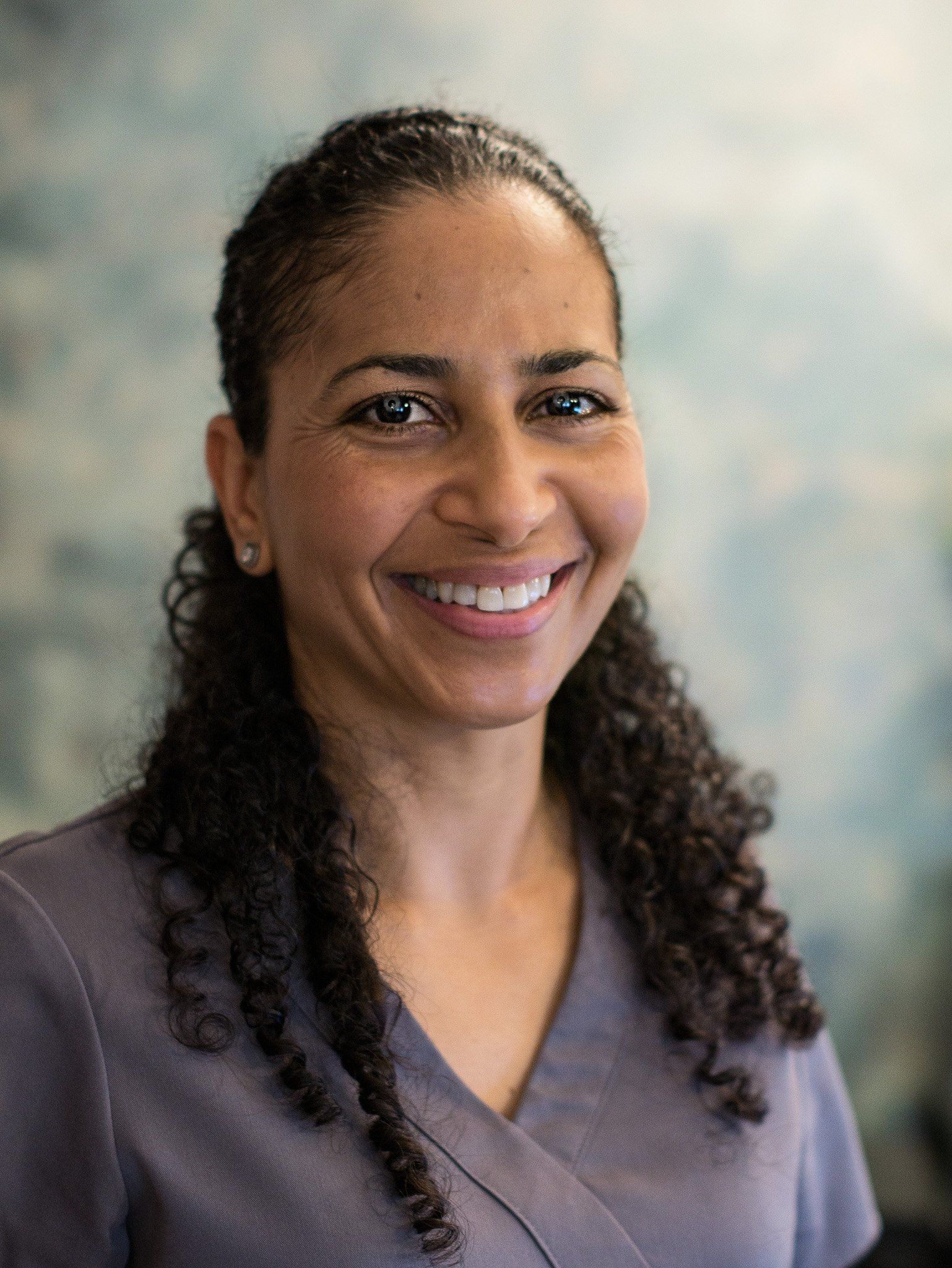 A woman with curly hair is smiling for the camera while wearing a scrub top.