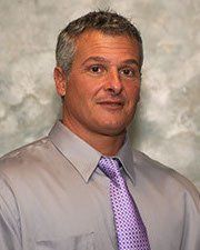 A man in a gray shirt and purple tie is standing in front of a wall.