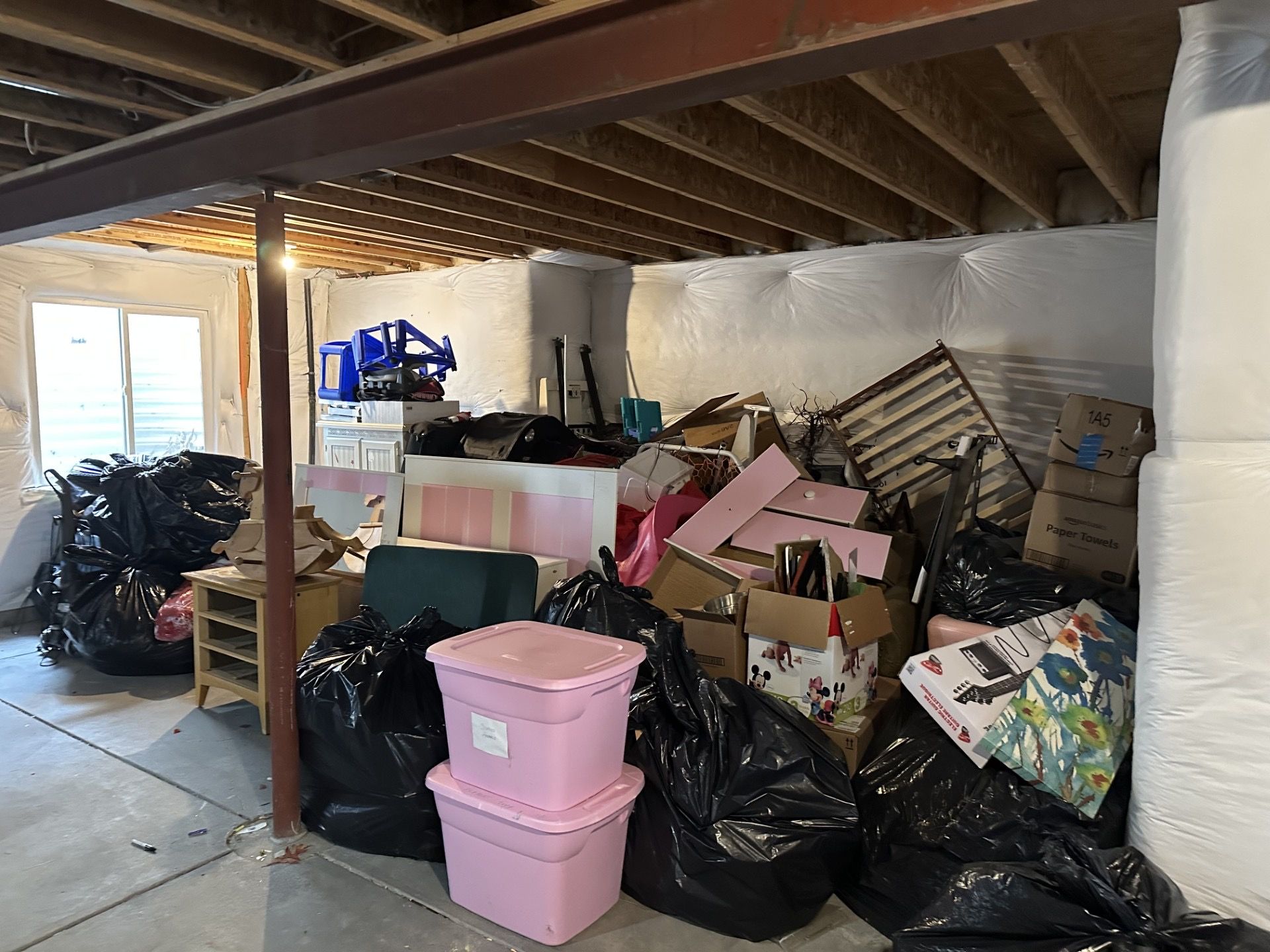 Basement storage area cluttered with black trash bags, boxes, and various items. Pink bins in front.