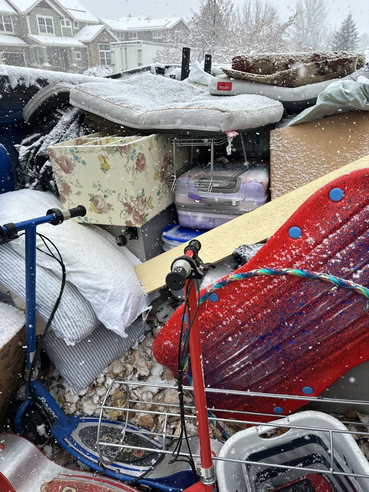 Pile of discarded household items in the snow; includes scooter, cushions, lamp, and a water cooler.