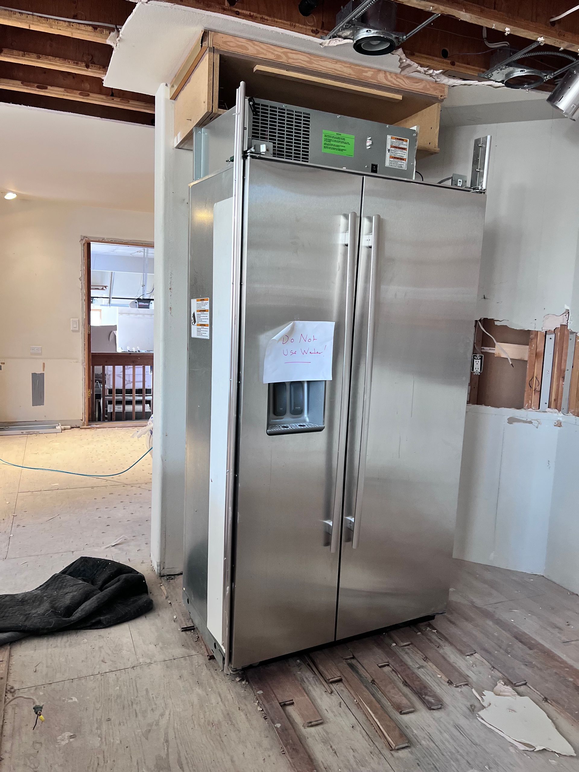 Stainless steel refrigerator in a room under construction, with exposed ceiling beams and unfinished flooring.