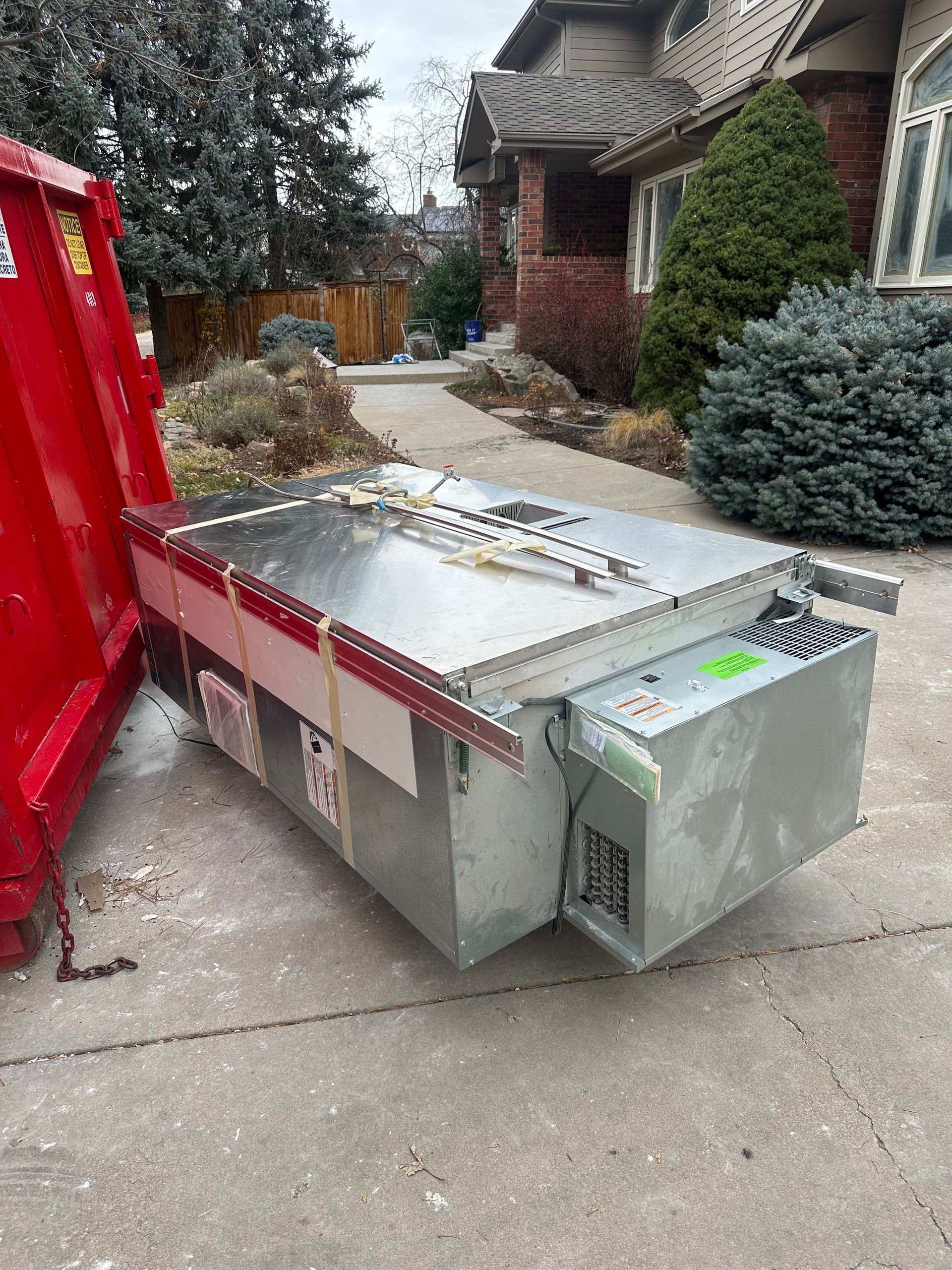 A large, industrial refrigeration unit sits on a driveway next to a red dumpster.