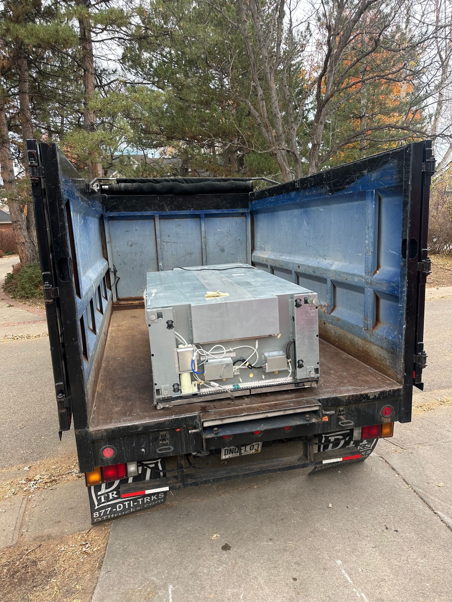 A large rectangular metal machine inside the bed of a blue and black dump truck, outdoors.