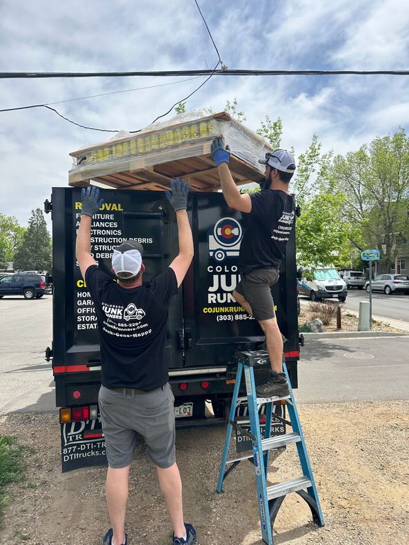 Two men loading a pallet into a the Colorado Junk Runners truck bed; one on a ladder. Outdoors, sunny day in Louisville, CO.