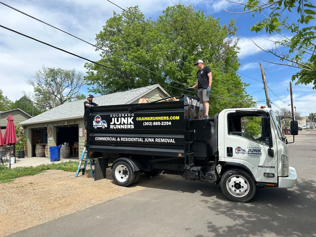Two men loading debris into a black and white Colorado Junk Runners truck on a street at a brewery in Louisville, CO.