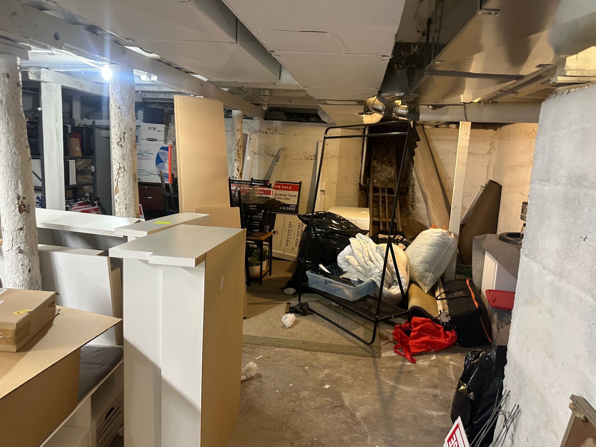 Cluttered basement with exposed pipes and concrete walls. Boxes, furniture, and trash are scattered throughout.