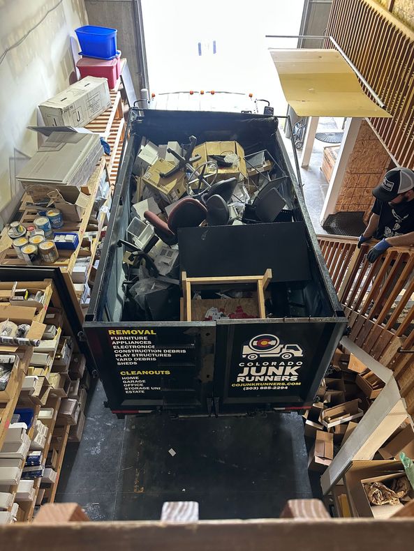 Colorado Junk Runners truck filled with debris in narrow space. Brick walls and stairs visible, possibly interior.