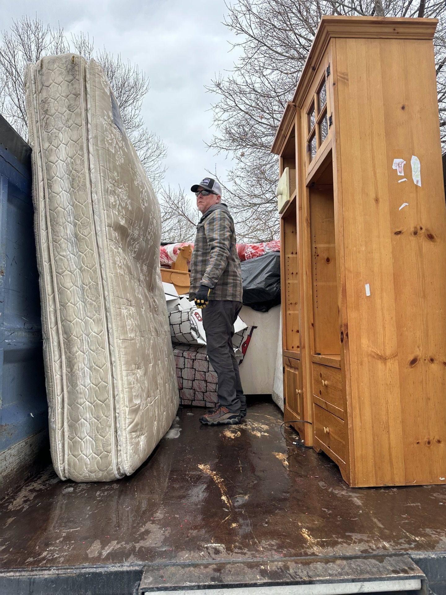 Man loading a mattress and cabinet into a truck. Cloudy day, outdoor setting.
