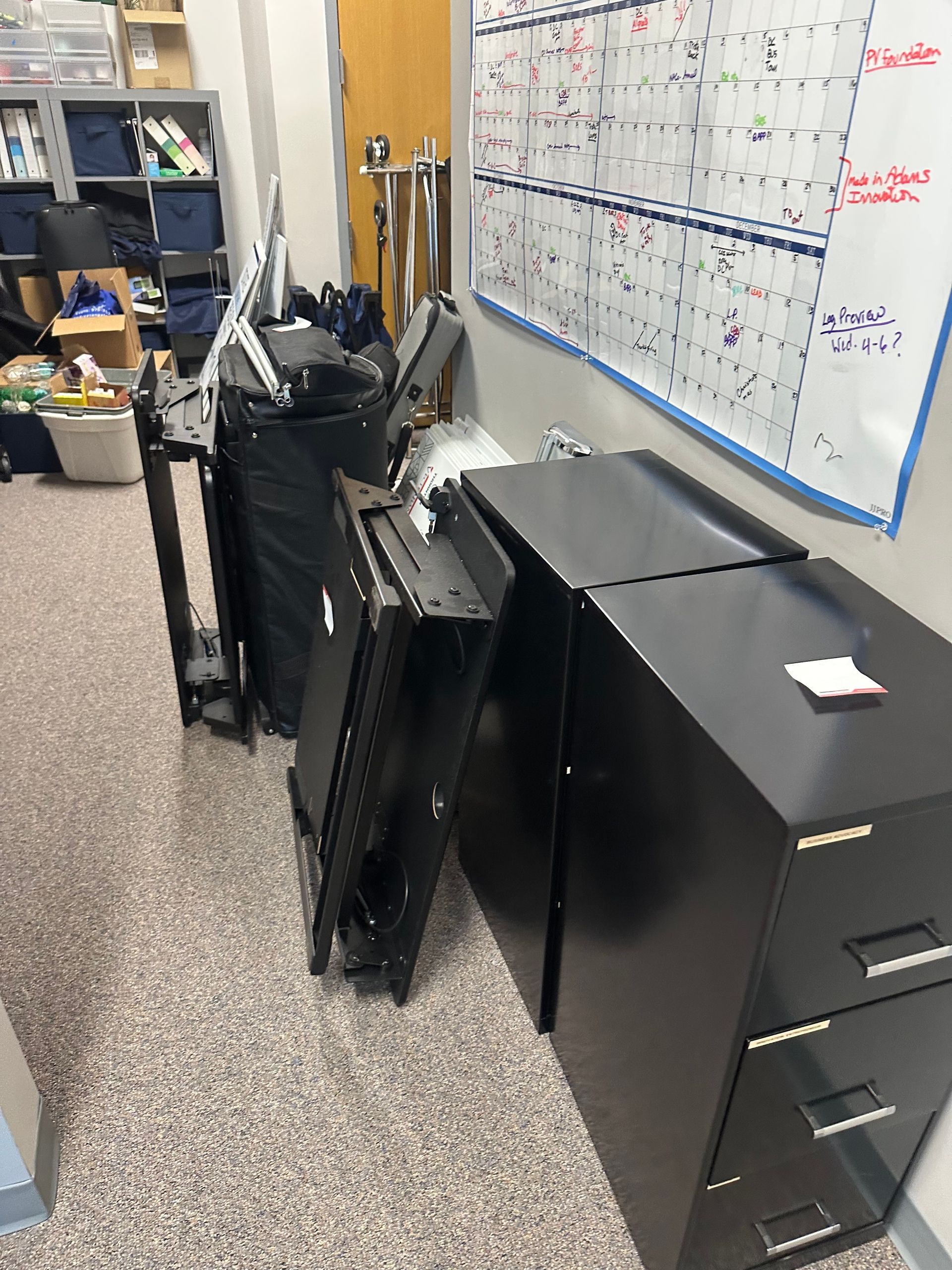 Black filing cabinet next to folded black metal shelves and a cart in a cluttered room.