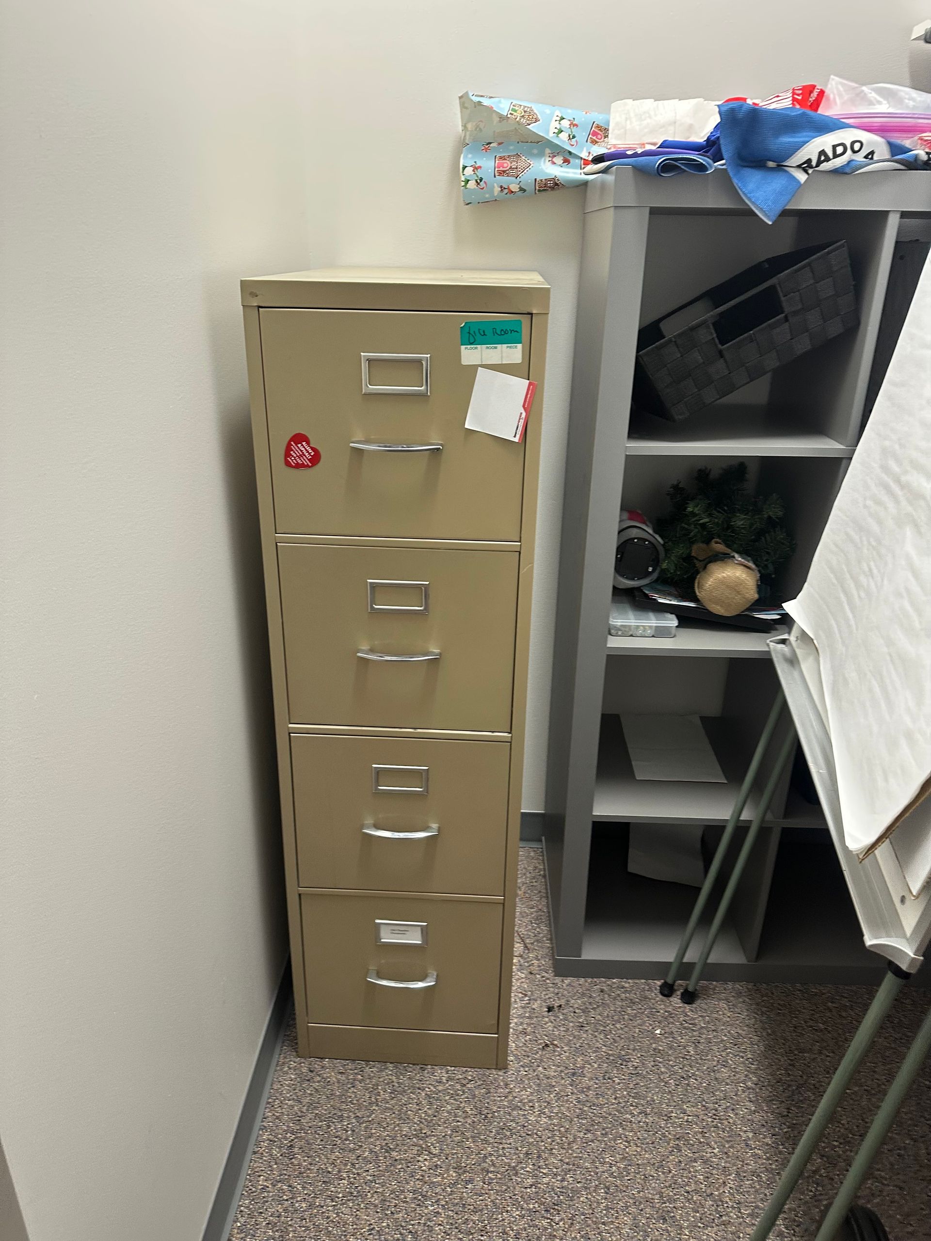 Tan file cabinet in a corner of a room. Next to an open shelving unit and a white board.