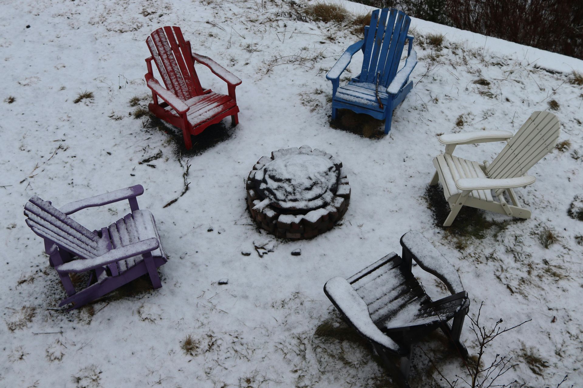 Five colorful Adirondack chairs in a snowy yard, arranged around a fire pit.