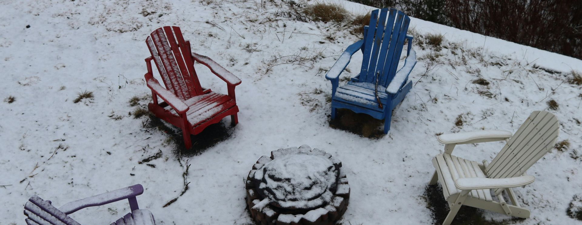 Five colorful Adirondack chairs circle a snow-covered fire pit.
