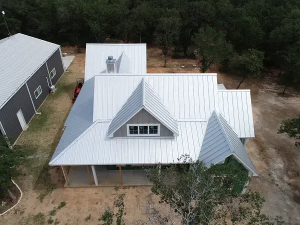 Aerial view of a white metal-roofed house surrounded by trees, with a dark outbuilding nearby