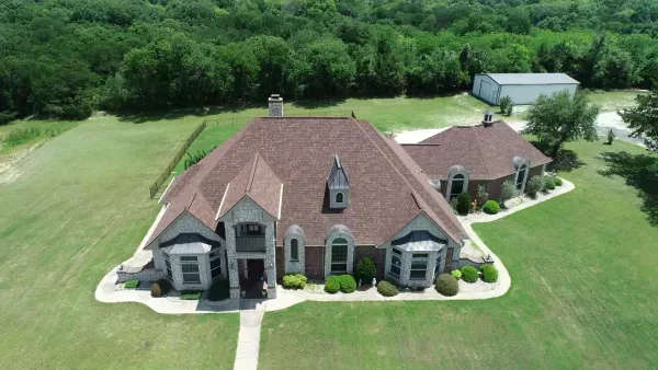 Aerial view of a large tan house with a red roof, surrounded by lawns and trees, with a driveway and outbuilding nearby