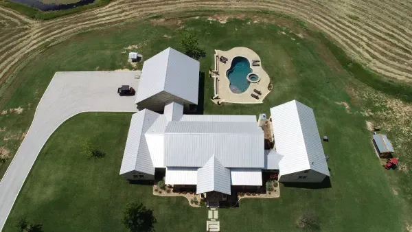 Aerial view of a white farmhouse with outbuildings, driveway, and backyard pool surrounded by green lawn.